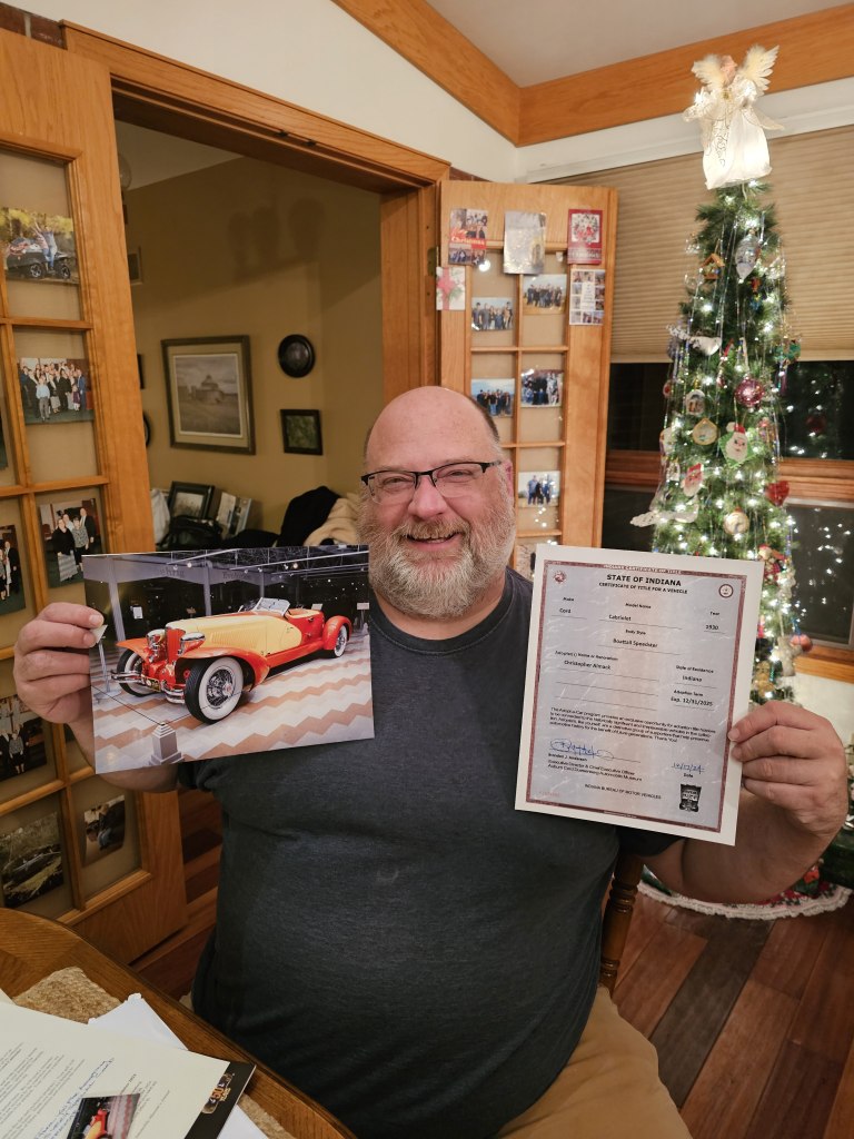 A man sitting at a table holding a photo of a classic car and an adoption certificate, with a Christmas tree in the background.
