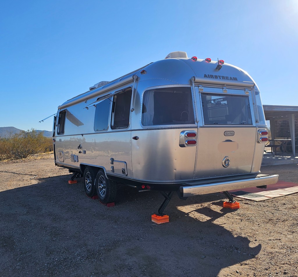 A shiny Airstream trailer parked in West Pinal County Park, with mountains in the background and clear blue sky.