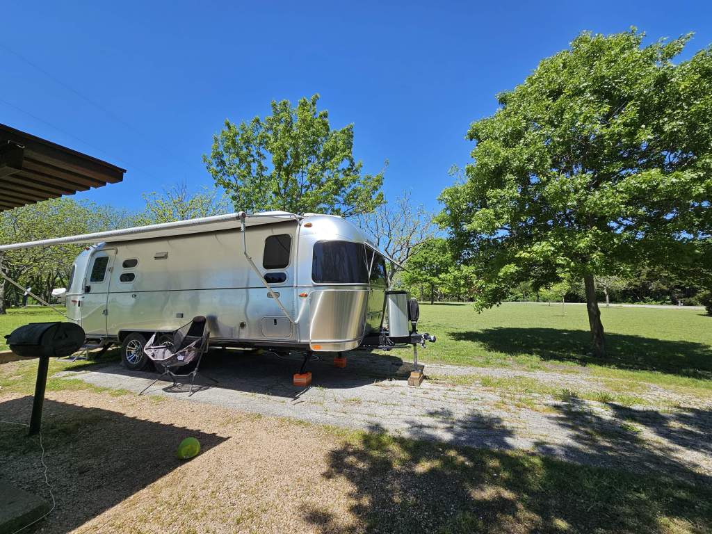 A silver Airstream trailer parked in a green, grassy area with trees and a clear blue sky.