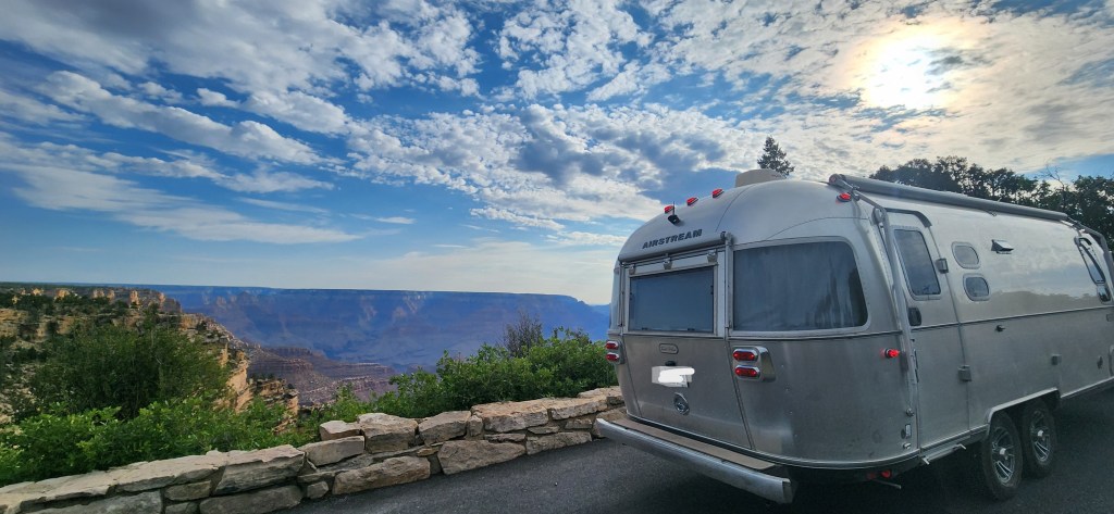 A silver Airstream trailer parked at a scenic overlook with a view of a canyon under a cloudy sky.