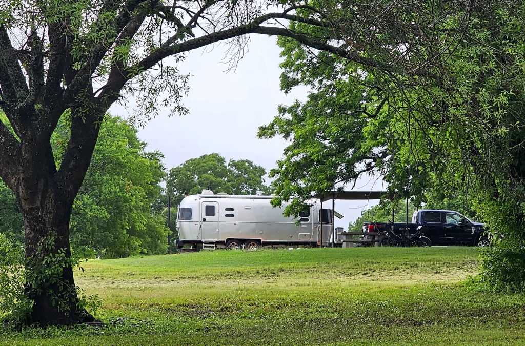 A silver Airstream trailer parked under a tree with a black pickup truck nearby, surrounded by green grass and trees.
